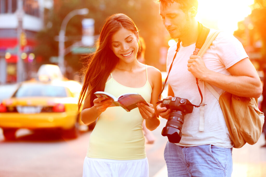 Travel tourist couple having a safe travel experience in New York reading guide book standing with SLR camera at sunset on Manhattan with yellow taxi cab in the background.