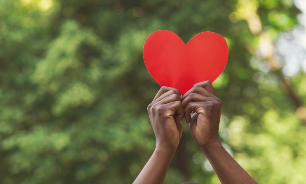 Love, charity and kindness concept. African-american millennial guy holding red paper heart on green nature background, empty space