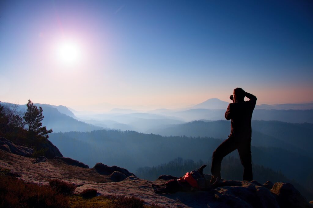 Professional photographer takes photos with mirror camera on peak of rock. Dreamy fogy landscape, spring orange pink misty sunrise in a beautiful valley below