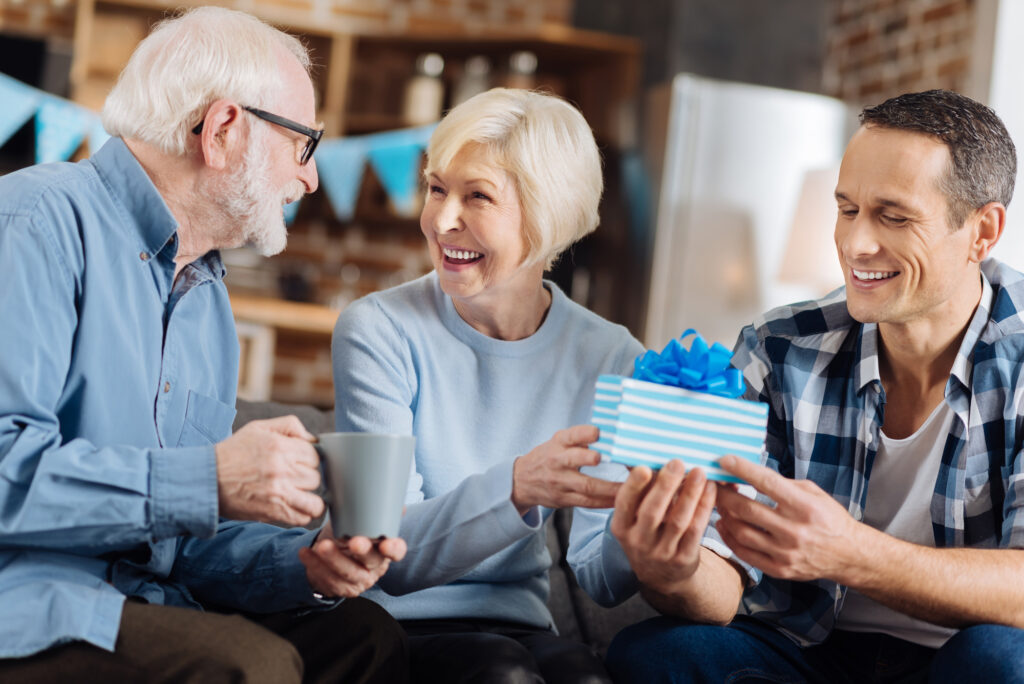 Happy birthday. Upbeat young man sitting on the sofa next to his elderly parents and giving a birthday present to his mother while she sharing happiness with her husband