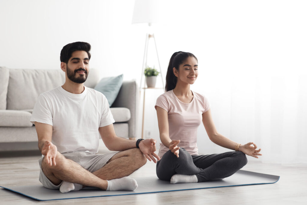 Happy couple doing exercises on quarantine at home, self-isolation together, stretching, exercise, health care at home. Young arab couple with closed eyes practices yoga, sitting on lotus at floor