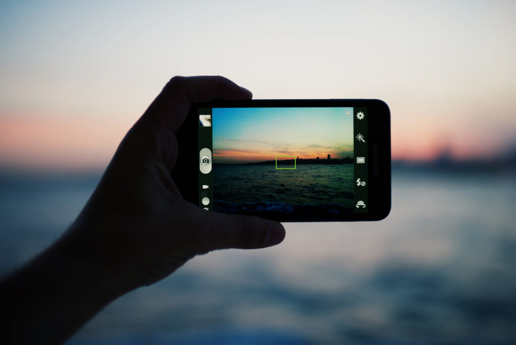 Person taking a photo of amazing sunset using smart phone camera, tourist hand holding cell phone while taking a photograph of nature landscape in travel,taking a picture of outdoors, blur background