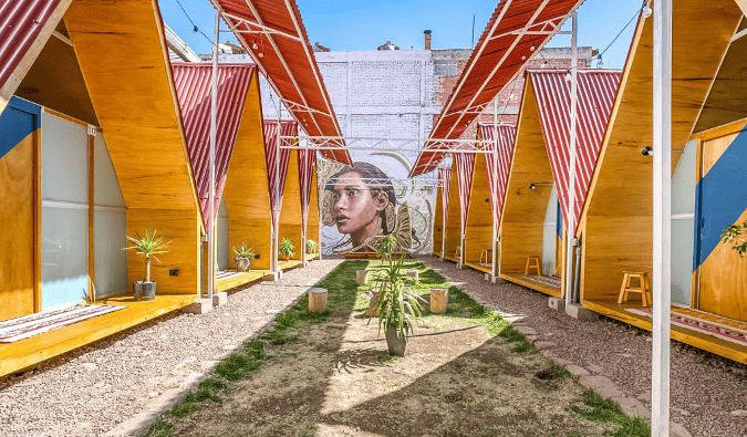 Individual A-frame houses lined up in two rows with green space and a large mural in the middle at Selina Plaza de Armas Cusco, a hostel in Cusco, Peru